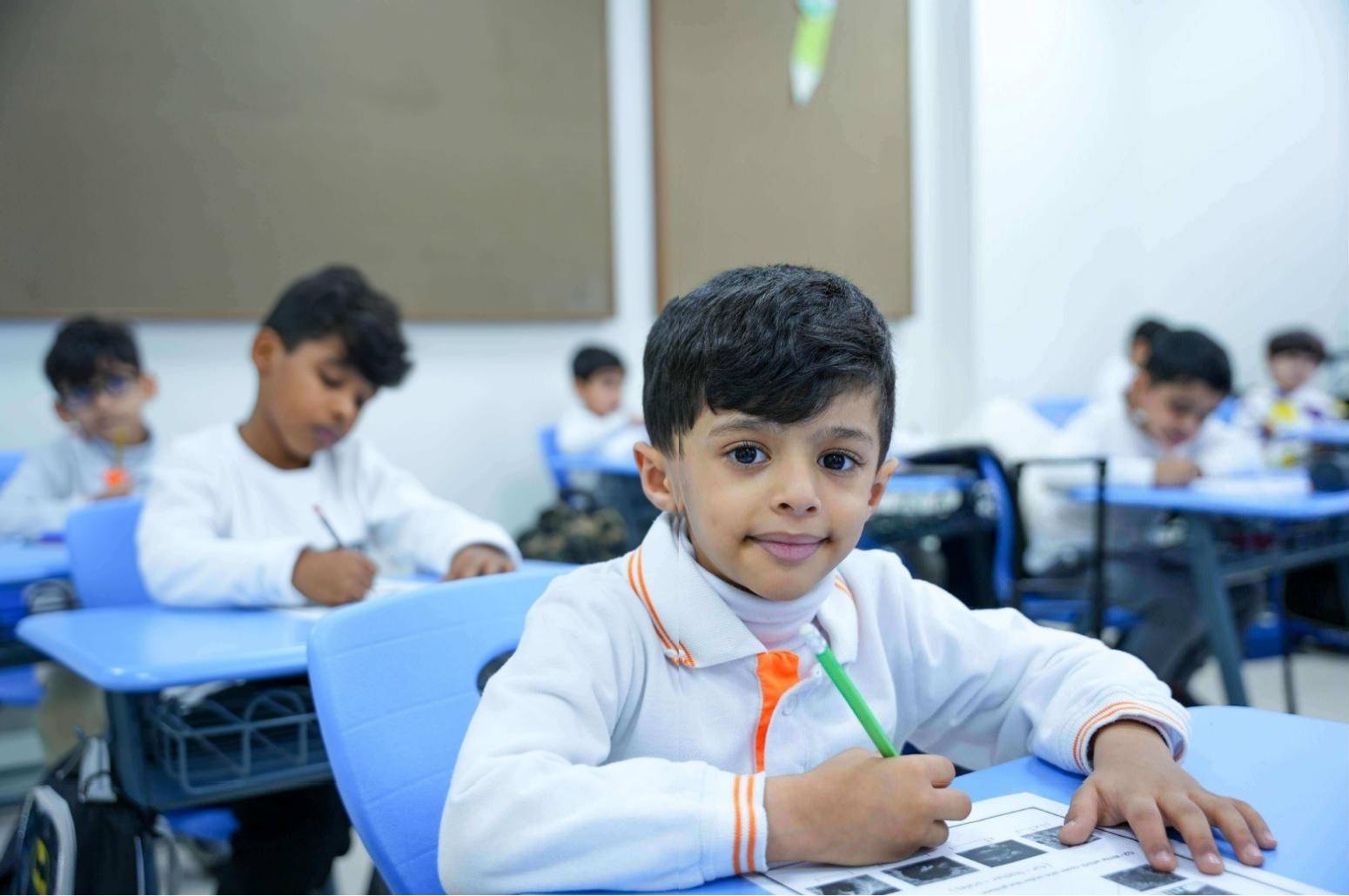 A BLS kindergarten student in the white polo with orange trim, smiling at his desk